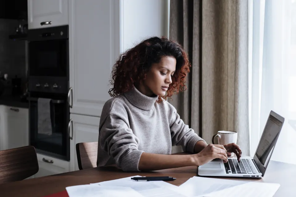 Mulher trabalha no notebook na mesa da cozinha, com papéis ao lado